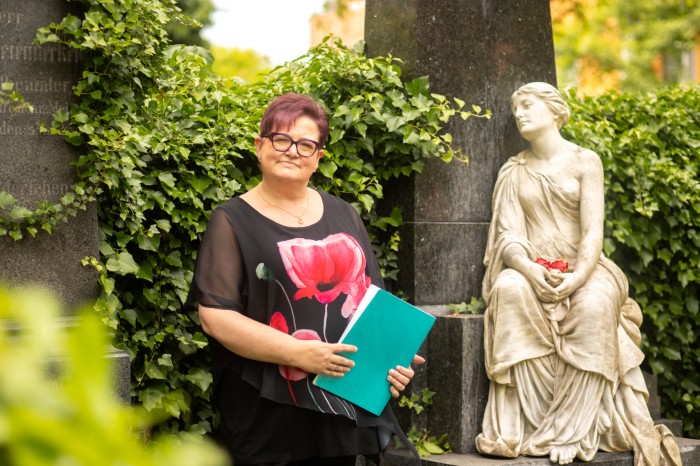 Steffi als Trauerrednerin neben einer Statue auf dem Friedhof.